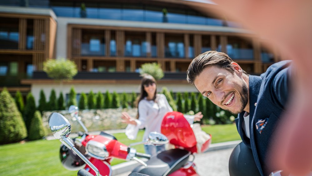 Le foto più belle dell’Alpen Tesitin Uomo che fa un selfie davanti a due scooter con una donna sullo sfondo