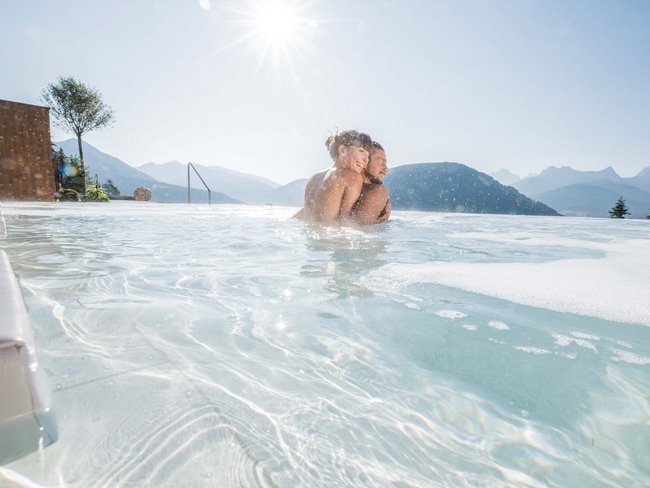 Your hotel in Val Pusteria/Pustertal with pool oasis Couple relaxing in outdoor pool with mountain view and sunshine