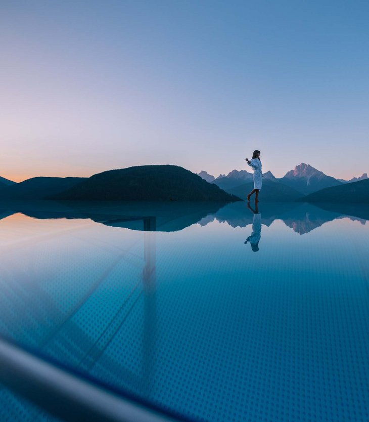 Ihr Hotel im Pustertal mit 5 Sternen: Alpen Tesitin Frau steht am Rand eines Infinity-Pools mit Bergblick bei Sonnenuntergang
