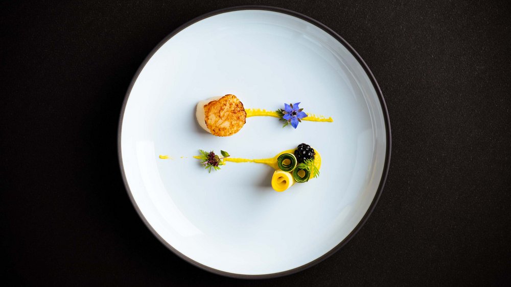 Le foto più belle dell’Alpen Tesitin Cappasanta impiattata artisticamente con salsa gialla e fiori commestibili su piatto bianco