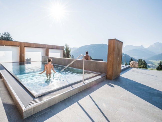 Your hotel in Val Pusteria/Pustertal with pool oasis People relaxing in outdoor pool with mountain view on sunny day