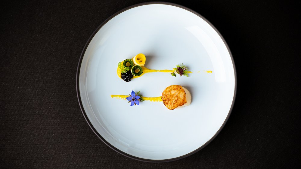 Le foto più belle dell’Alpen Tesitin Capesante artisticamente impiattate con rotoli di verdure e fiori edibili su piatto bianco
