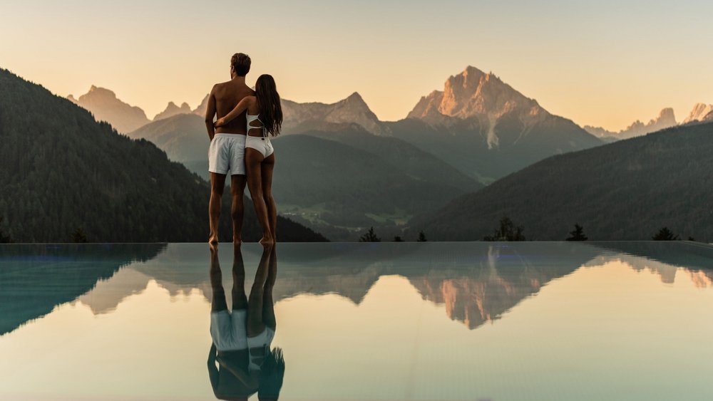 Le foto più belle dell’Alpen Tesitin Coppia in costume da bagno che guarda un paesaggio montano al tramonto, riflesso in piscina