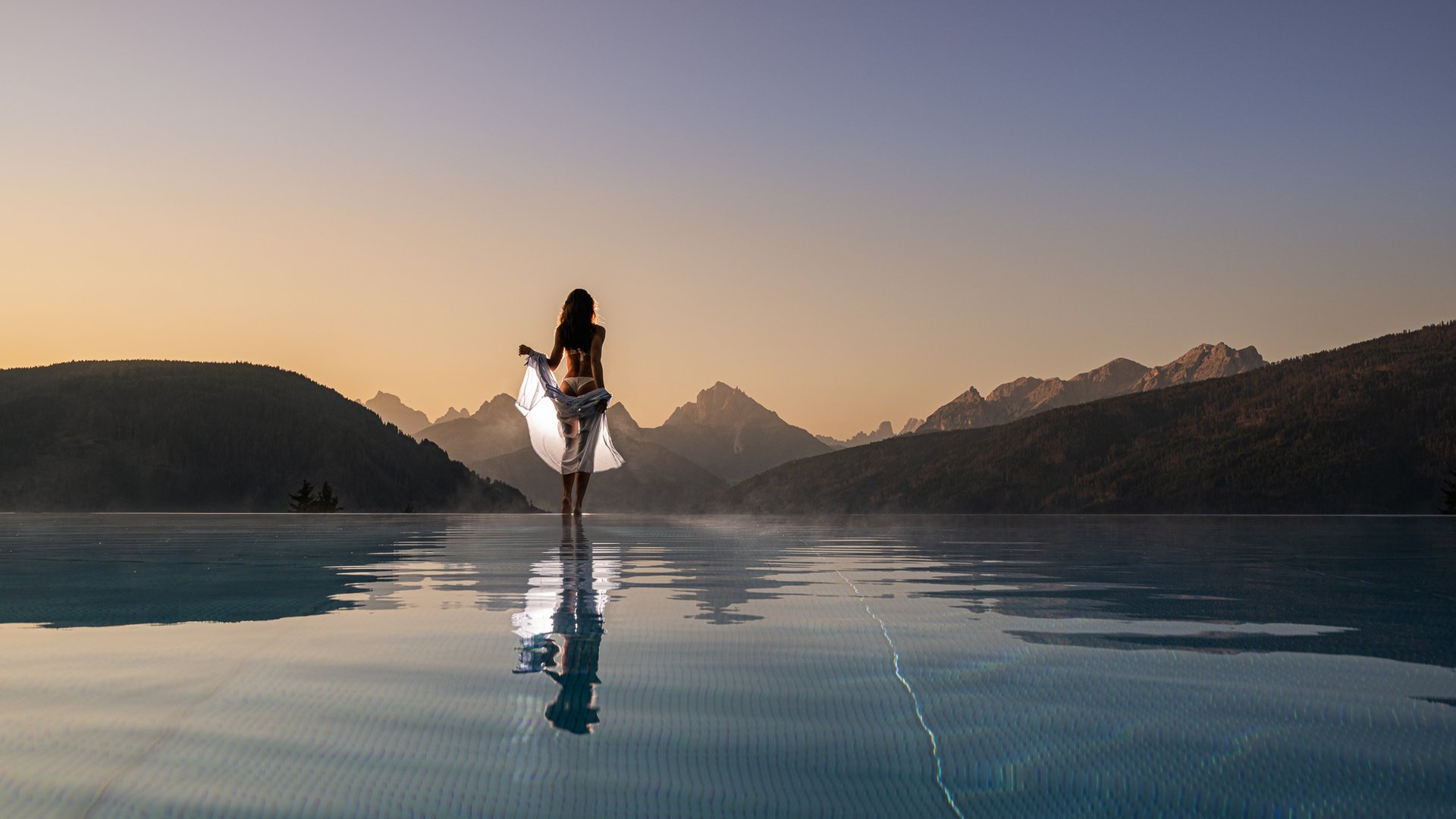 Ihr Hotel im Pustertal mit 5 Sternen: Alpen Tesitin Frau im weißen Tuch am Infinity-Pool mit Bergblick beim Sonnenuntergang