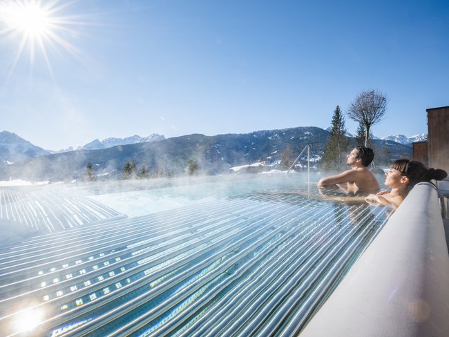 Your hotel in Val Pusteria/Pustertal with pool oasis Couple relaxing in heated pool with mountain view on a sunny winter day