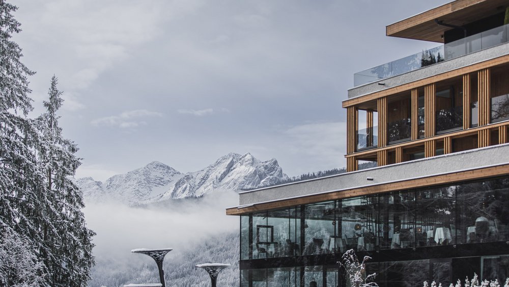 Le foto più belle dell’Alpen Tesitin Edificio moderno in vetro davanti a montagne innevate e pini in paesaggio invernale