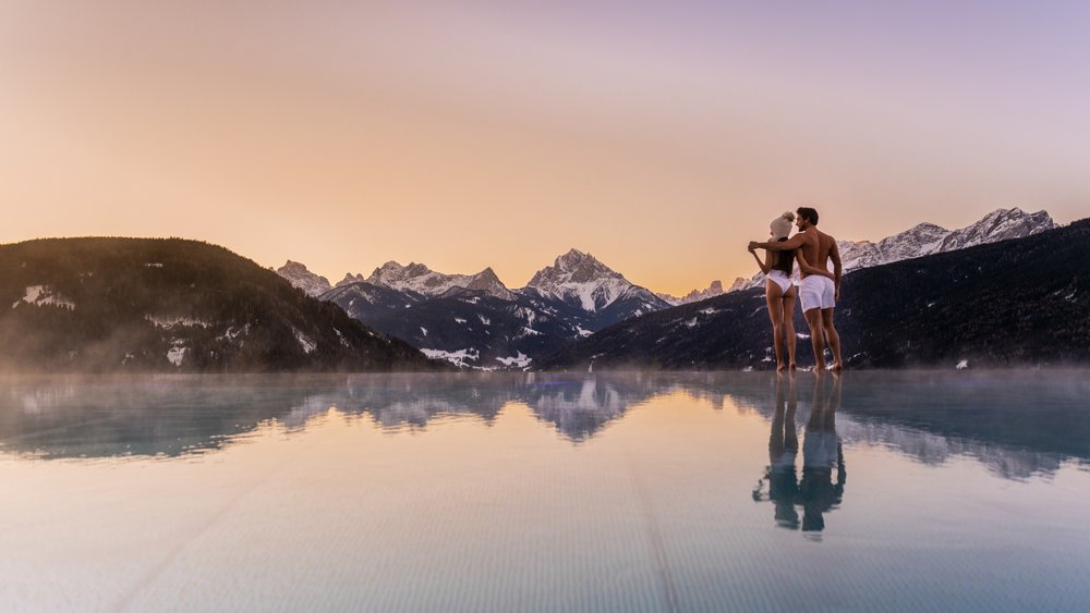 Le foto più belle dell’Alpen Tesitin Coppia in costume da bagno ai bordi di una piscina con montagne al tramonto