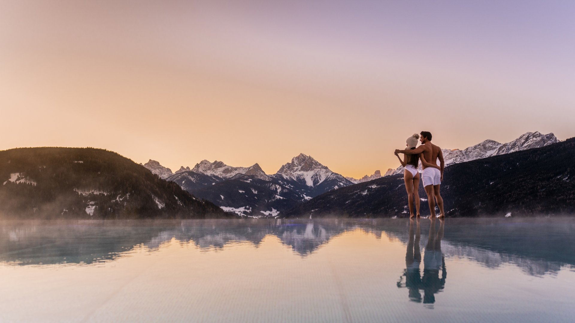 Ihr Hotel im Pustertal mit 5 Sternen: Alpen Tesitin Paar steht in Badebekleidung am Rand eines Pools vor Bergen im Sonnenuntergang