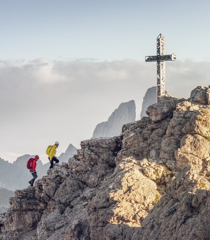Ihr Hotel im Pustertal mit 5 Sternen: Alpen Tesitin Zwei Bergsteiger besteigen felsigen Gipfel mit Metallkreuz bei sonnigem Himmel