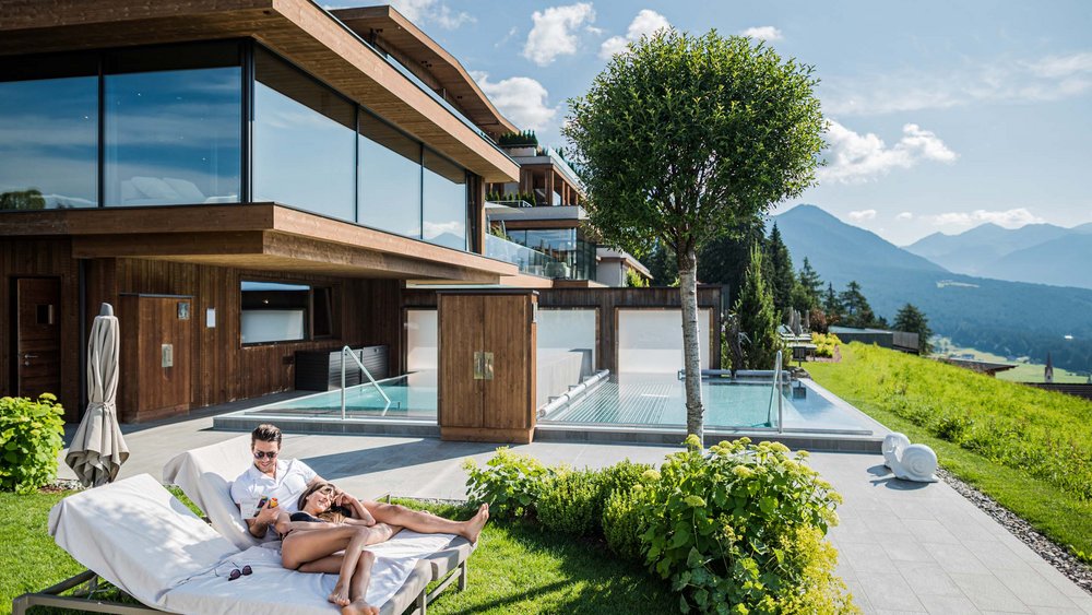 Le foto più belle dell’Alpen Tesitin Coppia rilassata su lettini davanti a casa in legno moderna con vista montagne e piscina