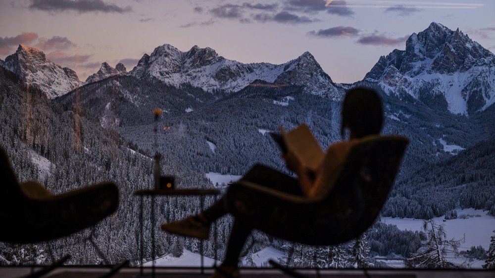 Le foto più belle dell’Alpen Tesitin Persona legge su una poltrona davanti a una grande finestra con vista sulle montagne innevate