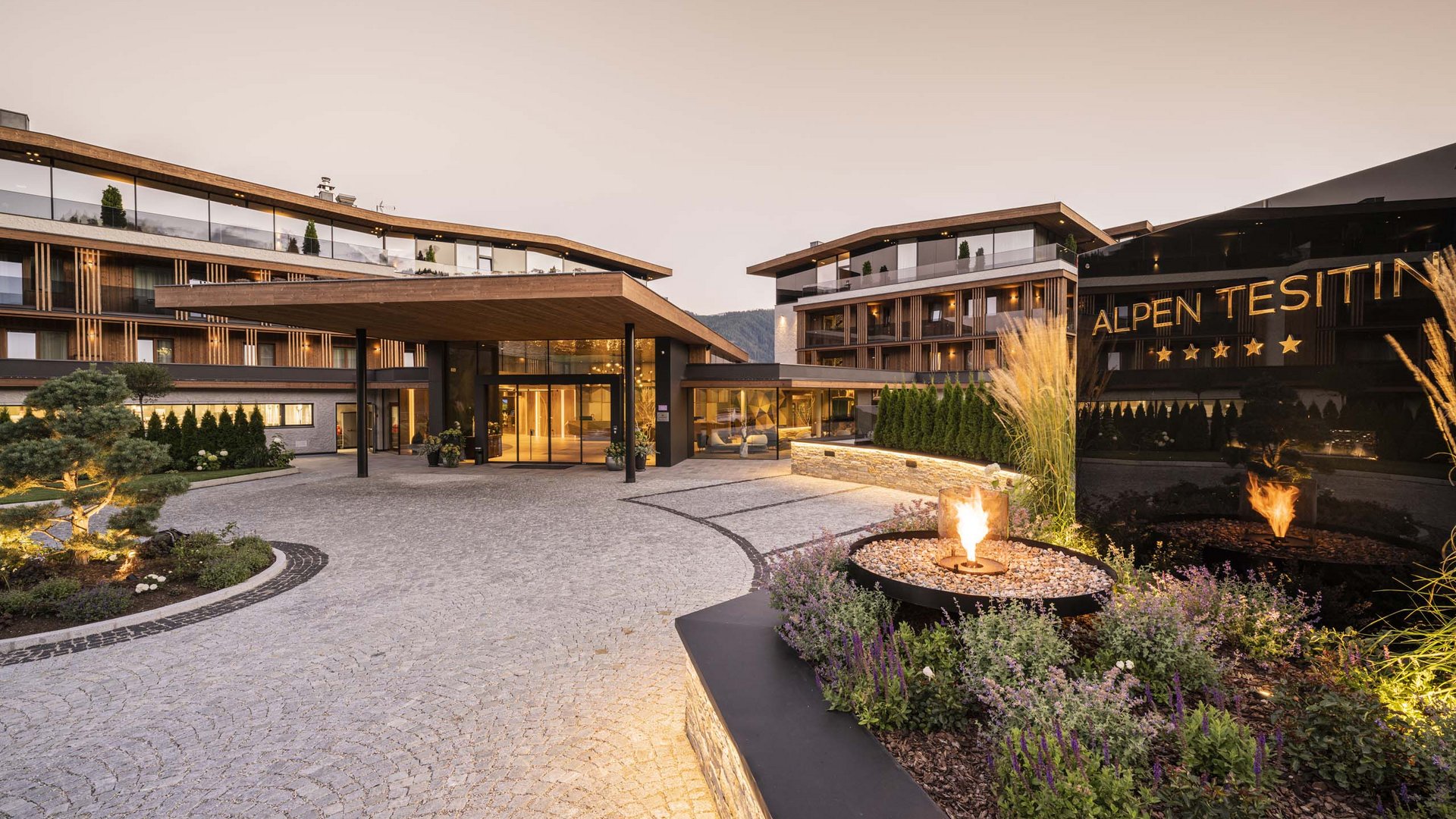 Ihr Hotel im Pustertal mit 5 Sternen: Alpen Tesitin Eingang eines modernen Hotels mit Feuerstelle und Alpenhintergrund bei Abenddämmerung