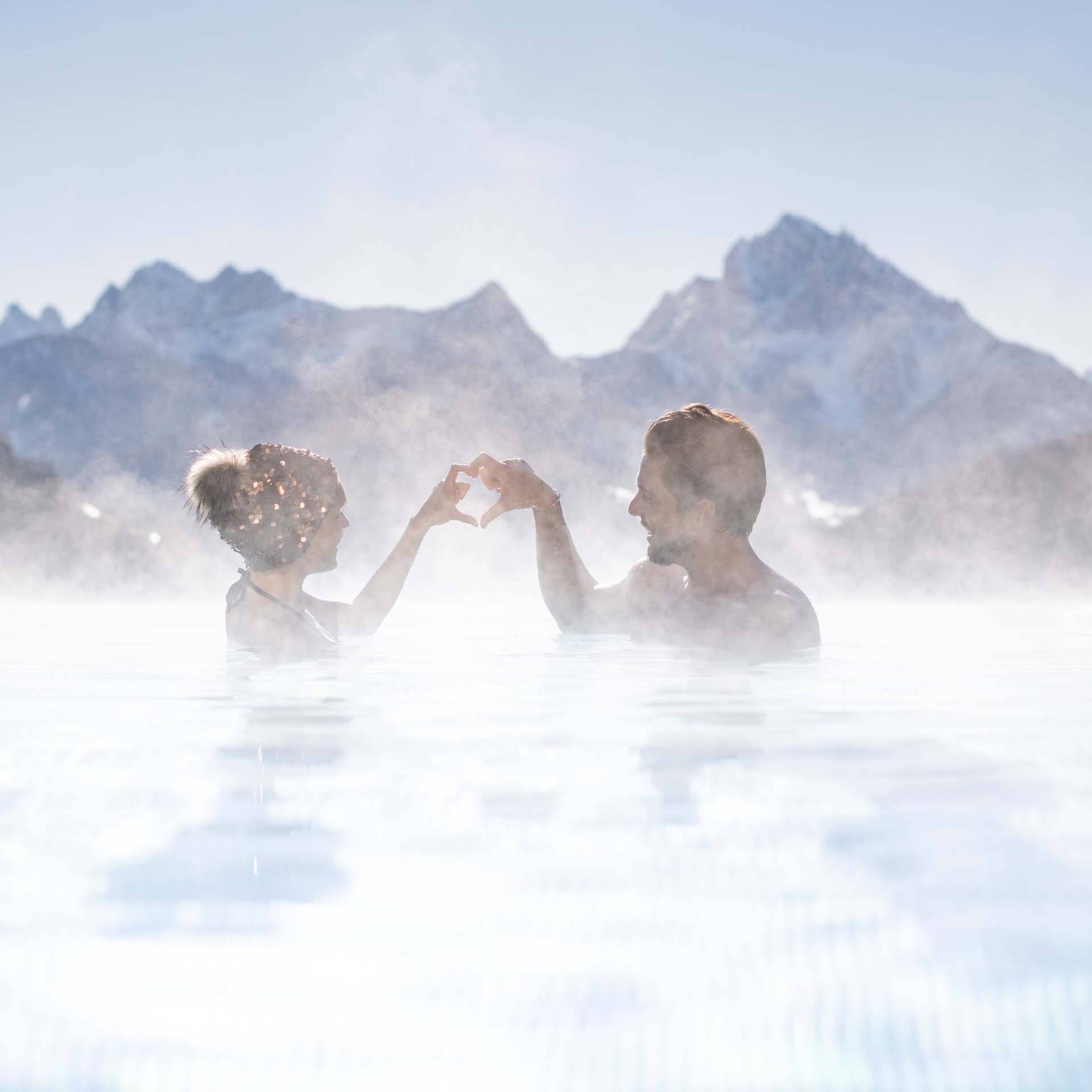 Ihr Hotel im Pustertal mit 5 Sternen: Alpen Tesitin Paar in heißem Pool vor Bergkulisse formt Herz mit Händen