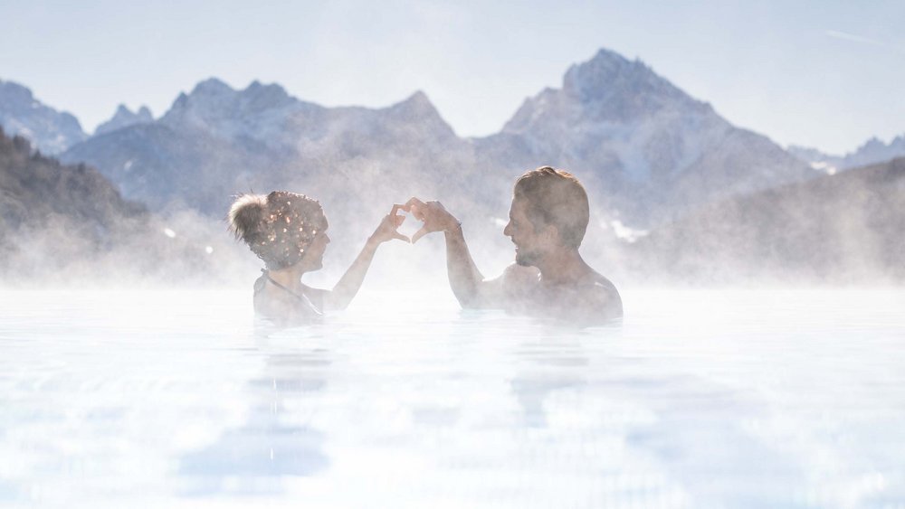 Le foto più belle dell’Alpen Tesitin Coppia in piscina calda con montagne sullo sfondo forma un cuore con le mani